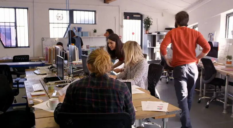 People in an office looking at computers; one person in orange sweater is walking around